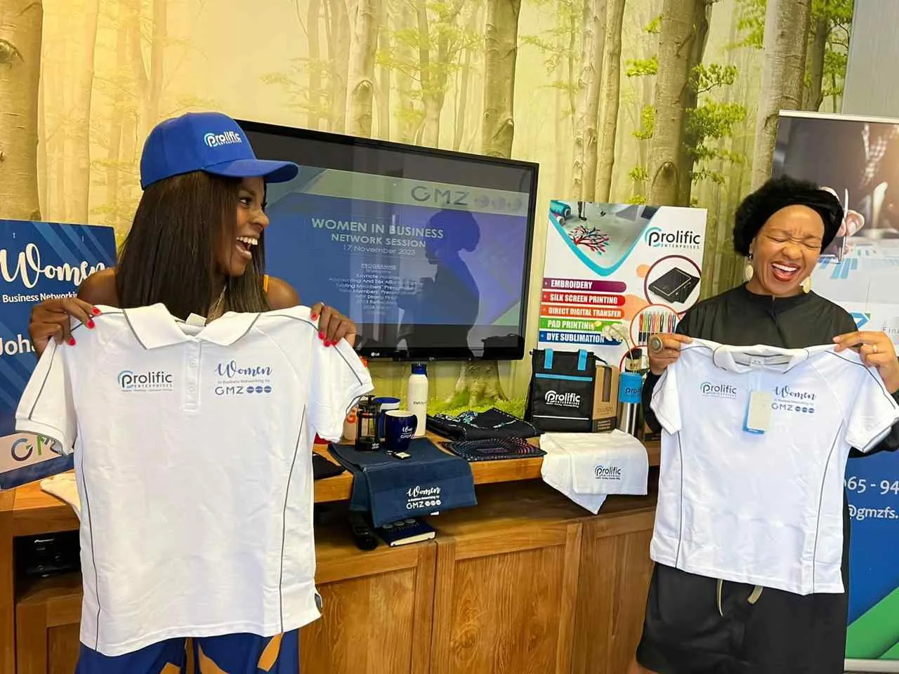 Two women at a Prolific Enterprises stand holding up white golf shirts branded for a Women in Business networking session, with branded caps, mugs, bottles and bags on the counter beside them.